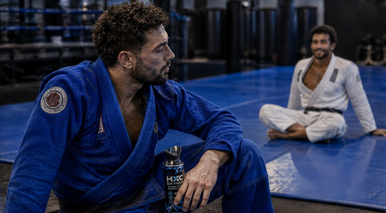 A jiu-jitsu athlete in a blue gi sits on a mat holding an HXC canister, with a training partner in a white gi behind him.